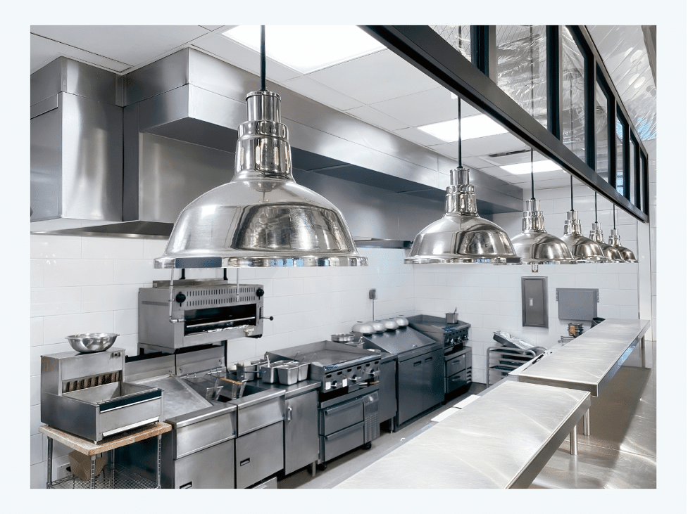 A professional commercial kitchen with stainless steel appliances, including stoves and ovens, stainless steel work surfaces, and industrial overhead lights. The space is clean and modern.