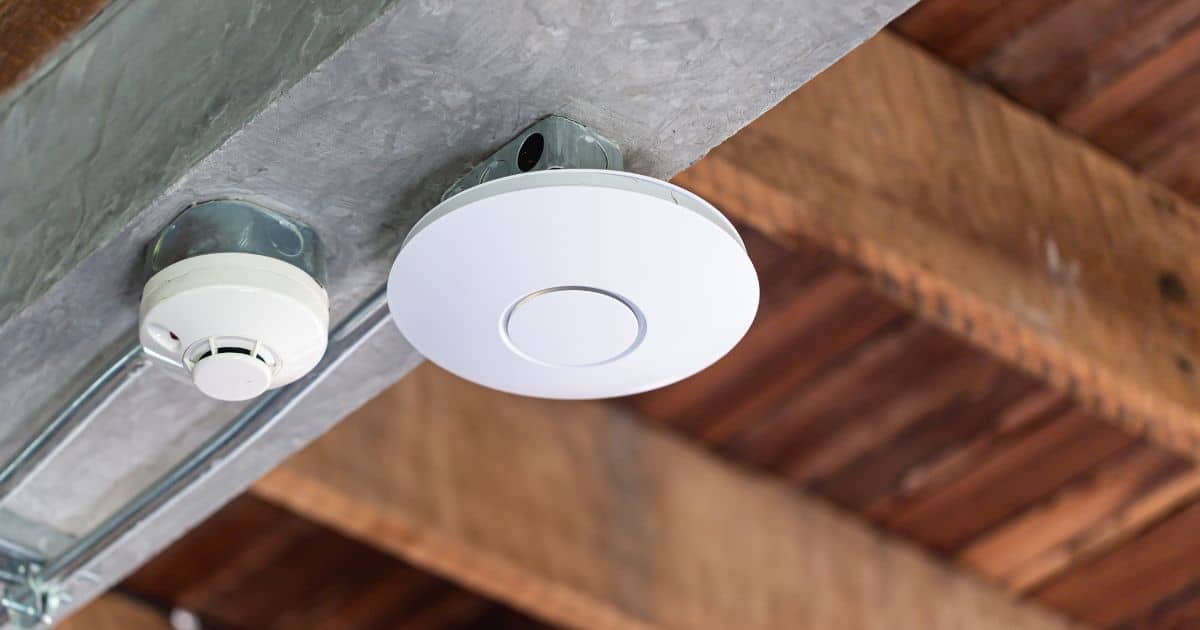 A white smoke detector and a circular ceiling-mounted device are attached to a metal beam with exposed wooden supports in the background.