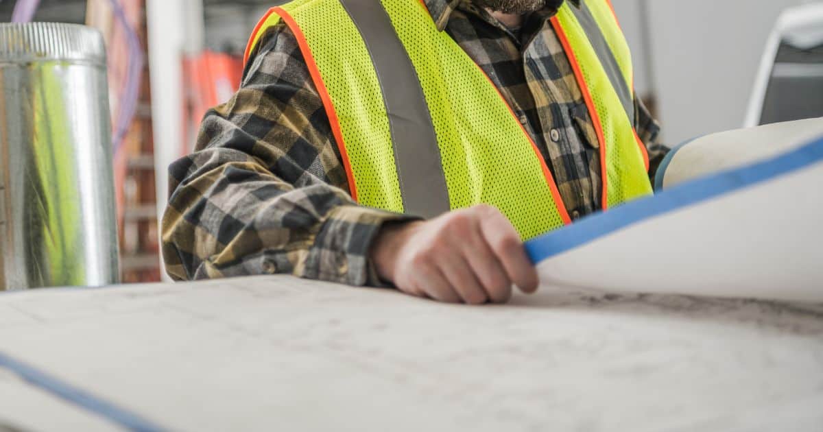 A person in a yellow safety vest examines a set of large blueprints or architectural plans.