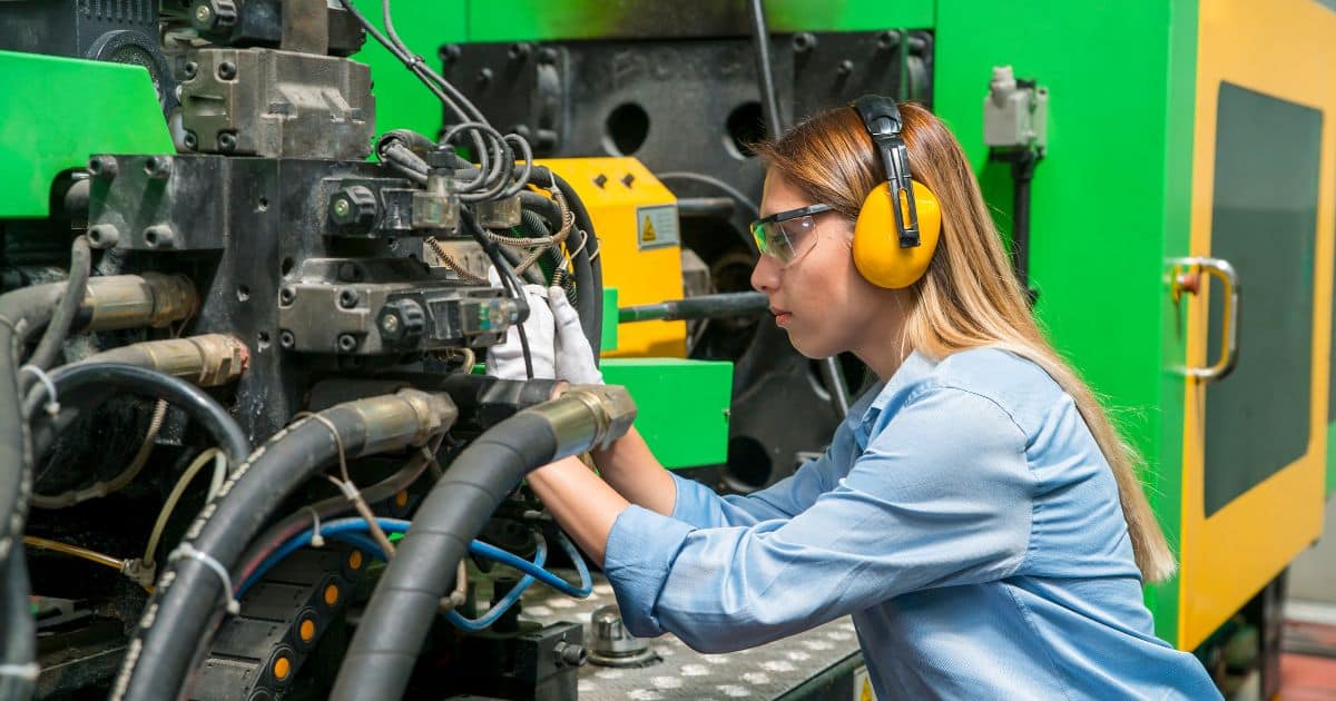 A female mechanical engineering technician wearing safety glasses and ear protection adjusts machinery in an industrial setting.