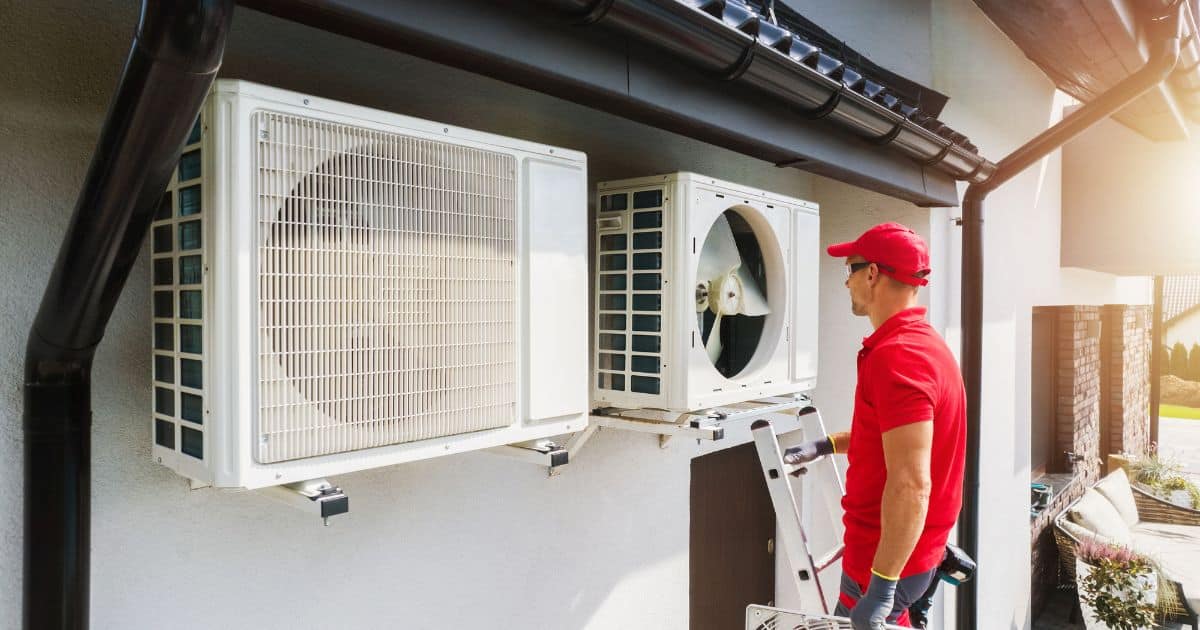 A heating and cooling systems technician in a red cap inspects an outdoor air conditioning unit attached to a house on a sunny day.