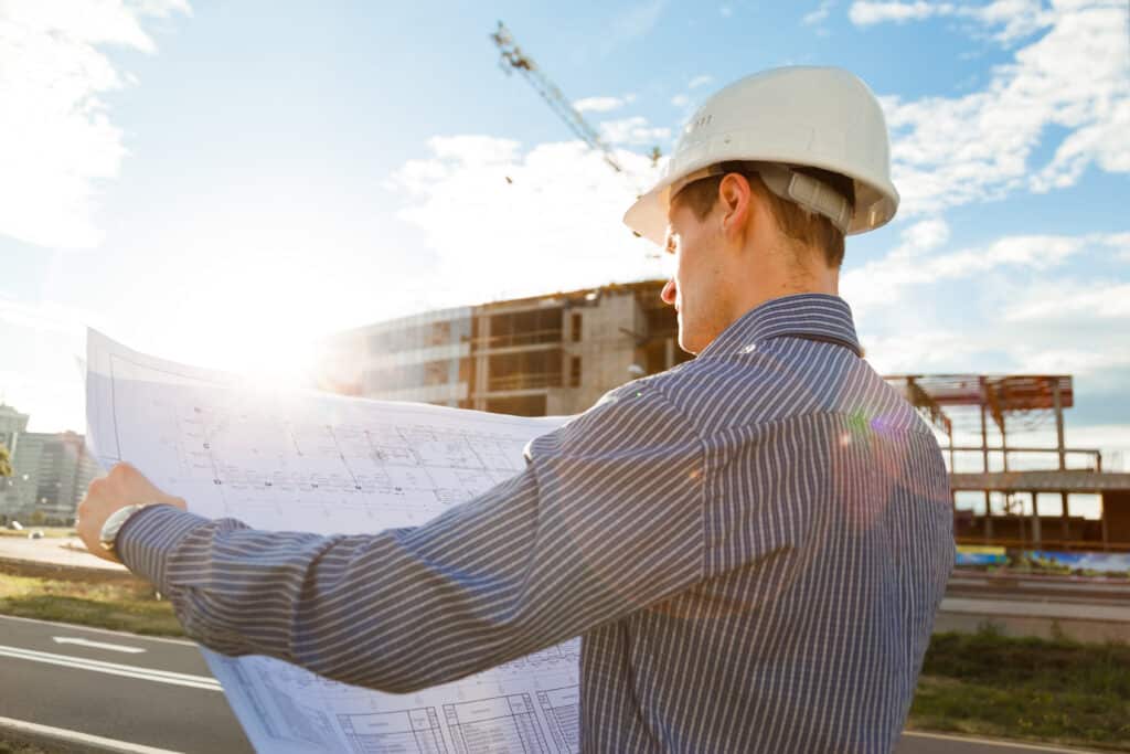 A man in a hard hat is holding a blueprint in front of a construction site.