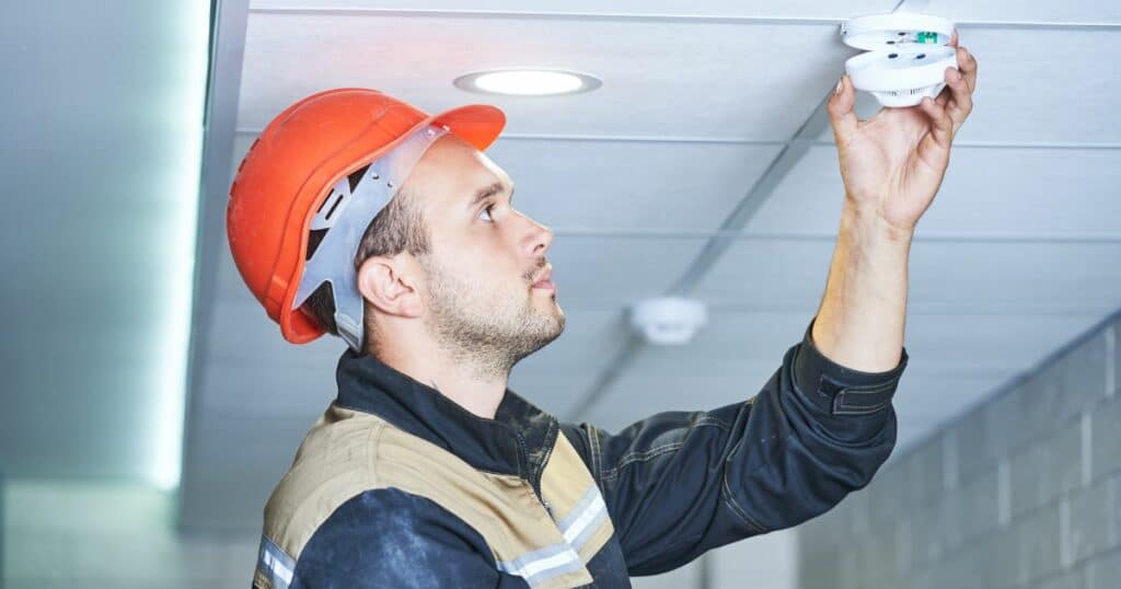 Electrician in hard hat installing a low voltage system smoke detector.