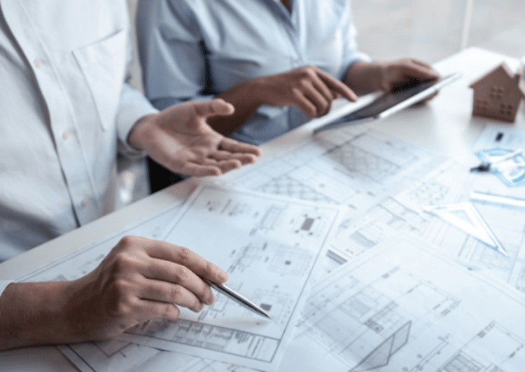 Two people review architectural blueprints on a table, one holding a pen and the other using a tablet, with a small house model nearby.