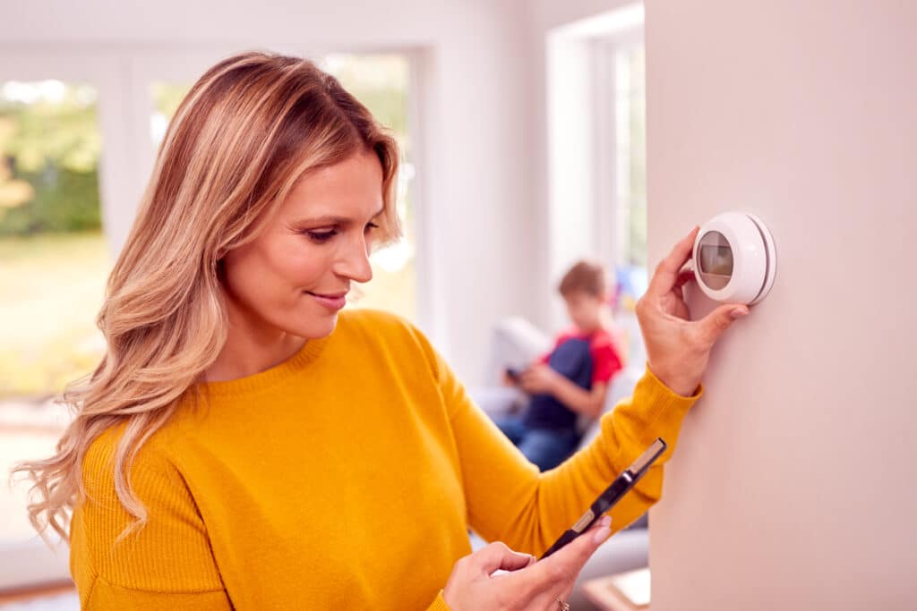 Woman in a yellow sweater adjusts a smart thermostat on the wall, holding a smartphone. A child is in the background, sitting and reading a book.