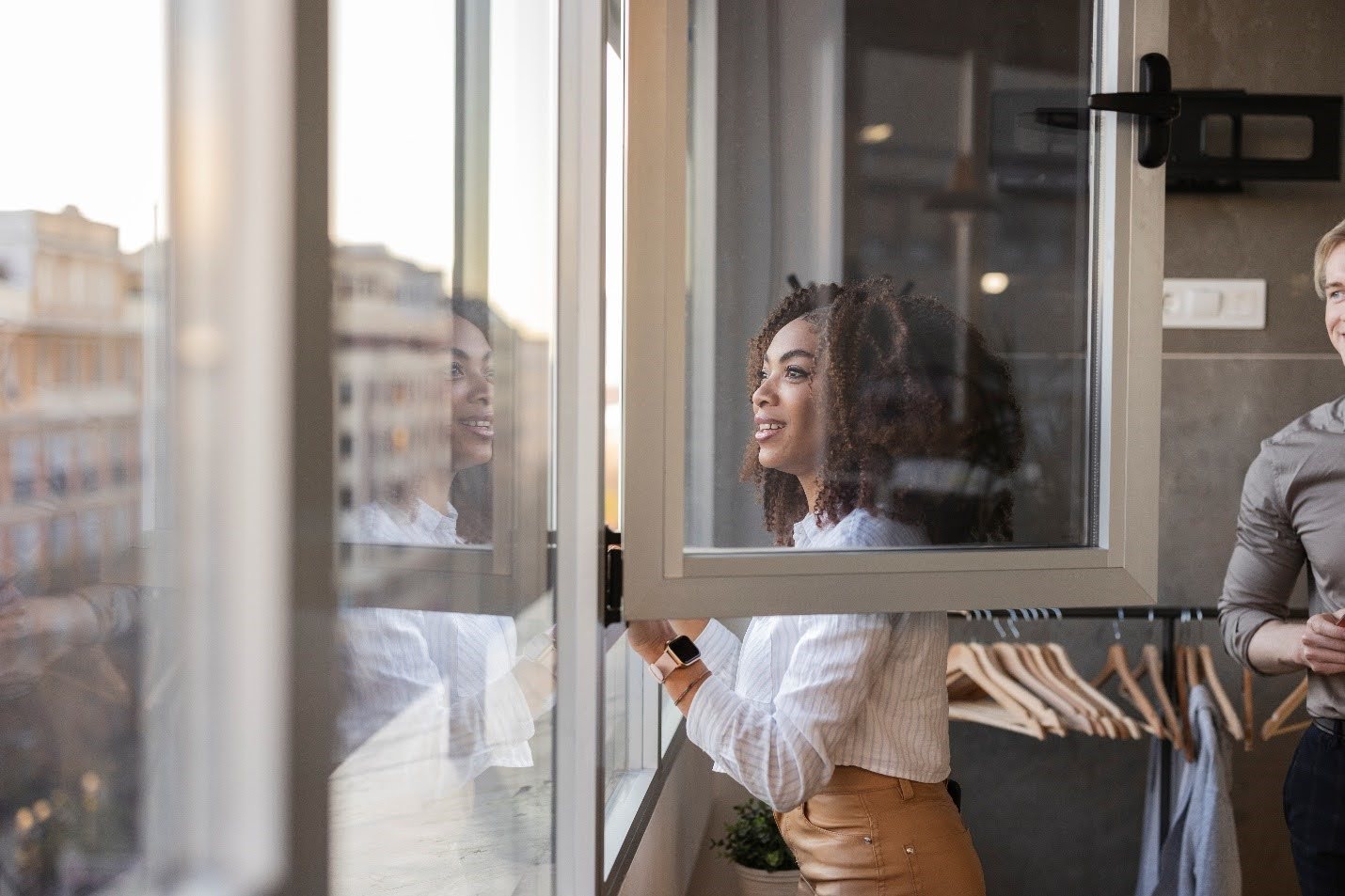 A woman in a white shirt stands by an open window, looking outside. She is reflected in the glass. A man in a gray shirt stands partially out of frame.