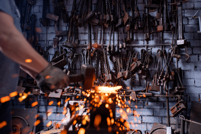 A person uses a hammer on hot metal, creating sparks in a workshop with various tools hanging on a wall in the background.