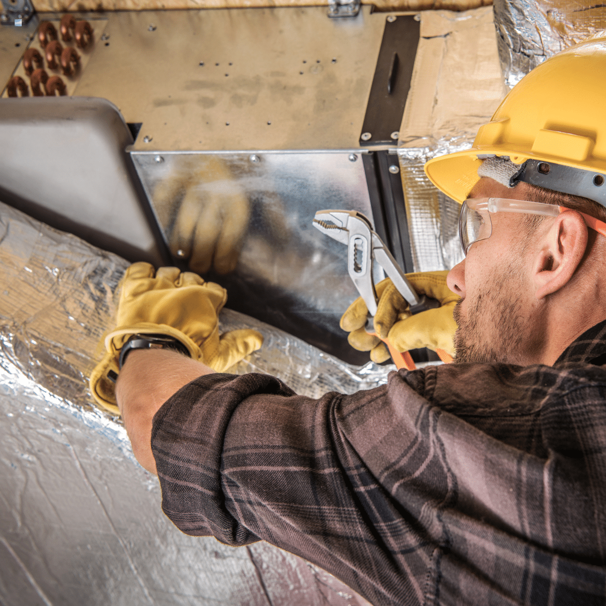 A worker wearing safety gear uses a wrench to adjust ductwork or HVAC equipment.