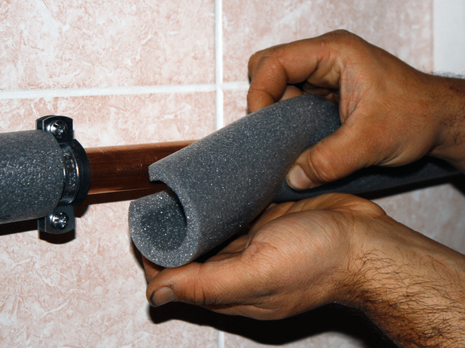 Hands installing foam pipe insulation around a copper pipe mounted on a tiled wall.