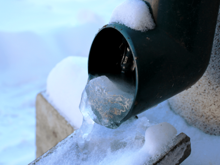 Close-up of a green metal downspout with bragging ice blocking the opening, surrounded by snow.