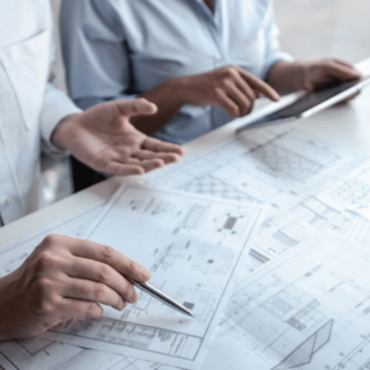 Two people review architectural blueprints on a table, one holding a pen and the other using a tablet, with a small house model nearby.